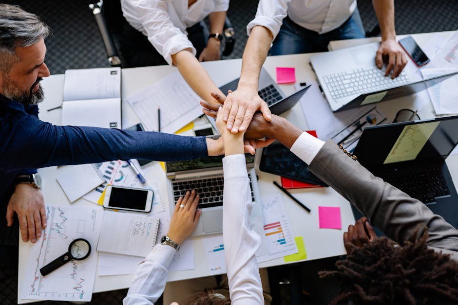 High-angle shot of diverse hands together over a desk with laptops and paperwork, symbolizing teamwork.